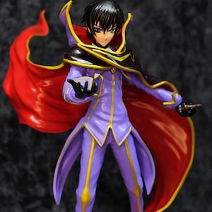 May include: A figurine of a character in a purple and gold outfit with a red cape. The figure has black hair and is holding a small object. The cape has a gold trim. The figure is standing in a dynamic pose.