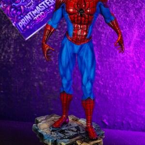 Puede incluir: Una detallada figura de Spider-Man con su cl&aacute;sico traje rojo y azul, de pie sobre una base rocosa. La figura tiene una pose din&aacute;mica, con un dise&ntilde;o de telara&ntilde;a en el pecho. Un letrero morado con el texto "PRINTMASTER 3DD" est&aacute; en el fondo.
