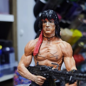 May include: A detailed figurine of a muscular man with black hair, a red headband, and a red scarf. The figurine is holding a black machine gun. The figure has a serious expression.