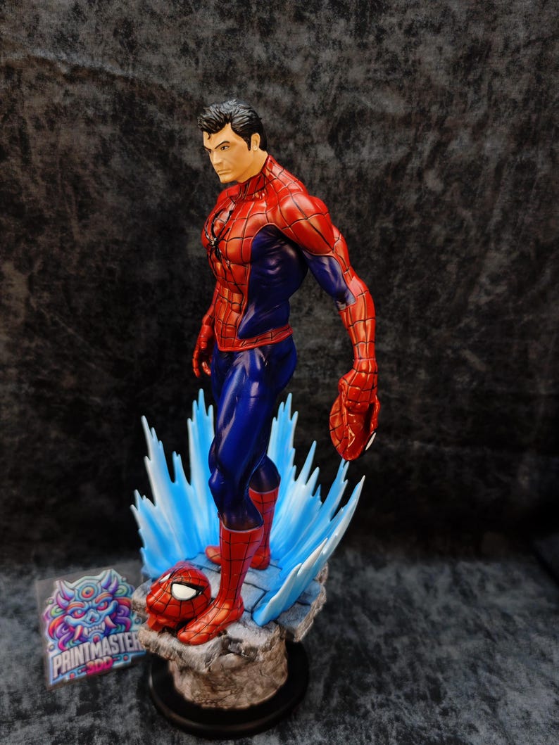 Op de afbeelding: Gedetailleerd beeldje van Spider-Man in een dynamische pose, met een rood en blauw kostuum. Het beeldje staat op een rotsachtige basis met blauwe energie-effecten en bevat een losgemaakt masker. Het Printmaster-logo is zichtbaar.