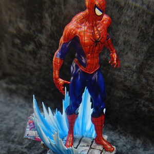 Op de afbeelding: Gedetailleerd beeldje van Spider-Man in een dynamische pose, gekleed in zijn klassieke rood-blauwe pak. Het beeld staat op een stenen voet met blauwe watereffecten. Het heeft een zwart spinnenembleem op de borst.