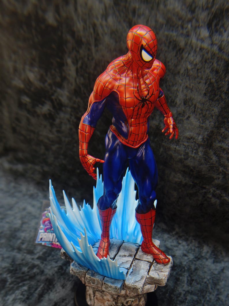 Op de afbeelding: Gedetailleerd beeldje van Spider-Man in een dynamische pose, gekleed in zijn klassieke rood-blauwe pak. Het beeld staat op een stenen voet met blauwe watereffecten. Het heeft een zwart spinnenembleem op de borst.