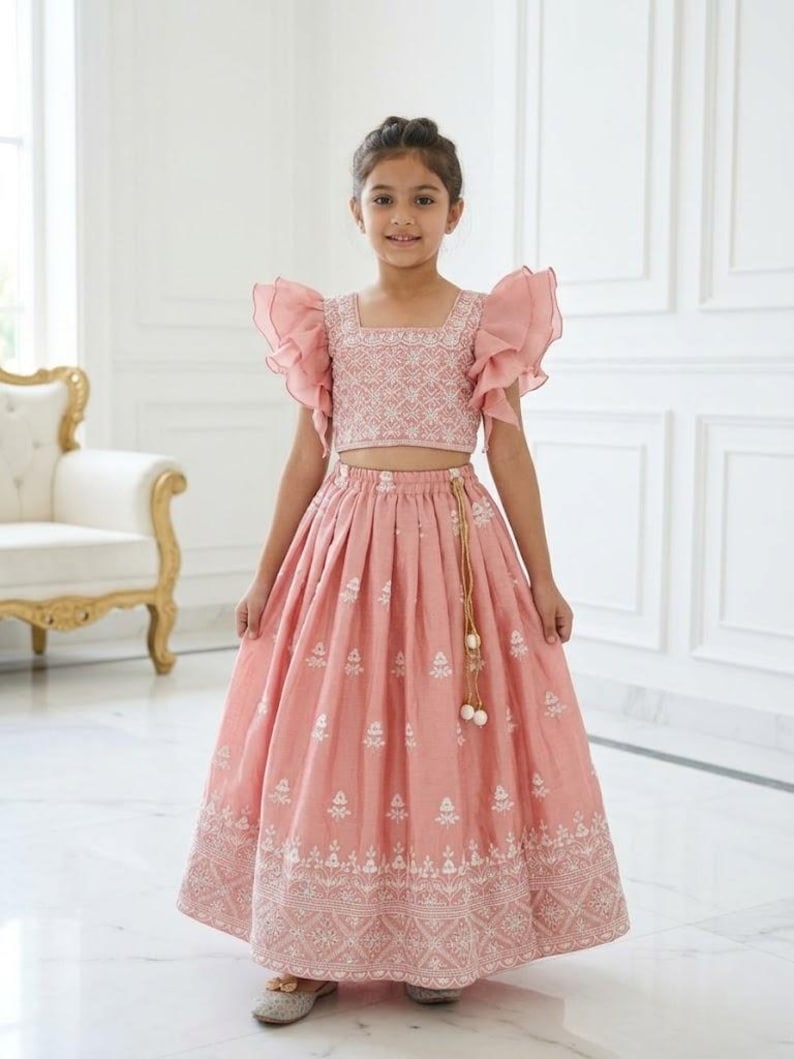 以下が含まれることがあります： ピンク色のツーピースの衣装を着た少女。トップスはスクエアネックとフリル袖。ロングスカートには白い刺繍が施されています。白いタッセルが付いたゴールドカラーのベルトがルックを完成させます。特別な機会に適しています。