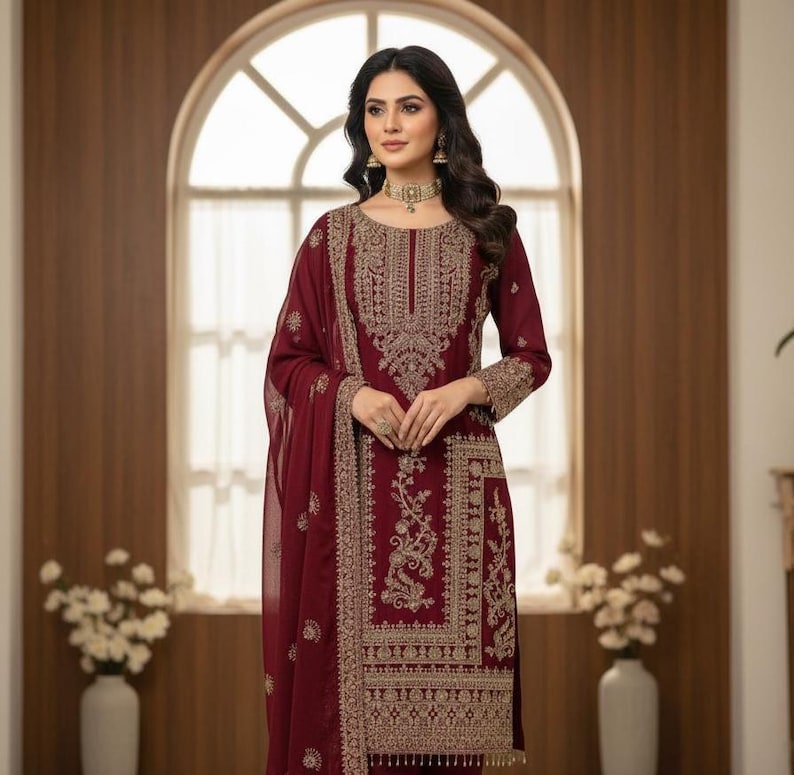 May include: A burgundy embroidered formal dress with a matching scarf. The dress features intricate gold threadwork around the neckline, sleeves, and hem. The outfit is worn with gold jewelry and is suitable for special occasions.