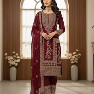 May include: Burgundy-colored embroidered salwar kameez ensemble. The outfit includes a long-sleeved tunic, matching pants, and a sheer scarf. The intricate gold embroidery adorns the neckline, sleeves, and pant cuffs. The outfit is worn with gold-colored heels.