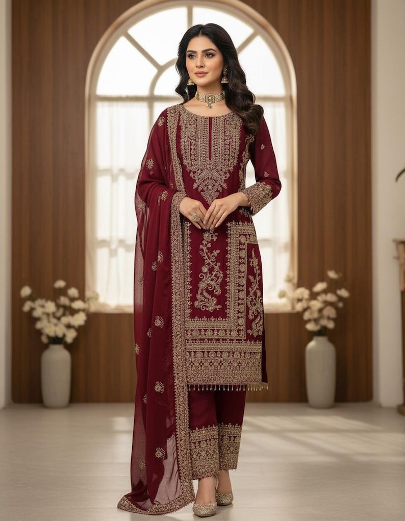 May include: Burgundy-colored embroidered salwar kameez ensemble. The outfit includes a long-sleeved tunic, matching pants, and a sheer scarf. The intricate gold embroidery adorns the neckline, sleeves, and pant cuffs. The outfit is worn with gold-colored heels.