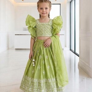 以下が含まれることがあります： 若い女の子がライムグリーンの衣装を着ています。 衣装には、フリル袖のクロップトップ、白い刺繍のロングスカート、お揃いのスカーフが含まれています。 衣装は伝統的なスタイルで、花のヘッドバンドとマッチングシューズが付いています。