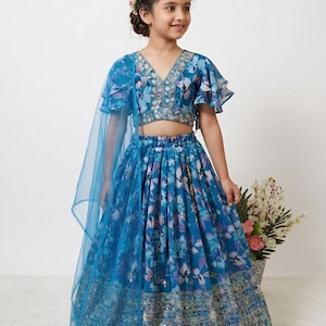 May include: A young girl wearing a blue floral print outfit. The outfit includes a crop top with ruffled sleeves, a long skirt, and a sheer blue scarf. The ensemble is adorned with intricate embroidery and embellishments.