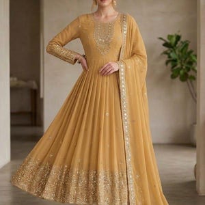 May include: A mustard yellow floor-length Anarkali dress featuring long sleeves, a round neckline, and intricate gold embroidery on the bodice, cuffs, and hem. The dress includes a matching sheer dupatta with a gold-trimmed border and mirror work.
