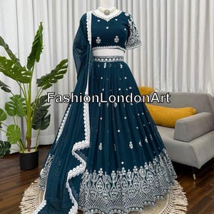 May include: A teal-colored formal dress with intricate white embroidery. The dress features a crop top, a flowing skirt, and a matching shawl. The text "FashionLondonArt" is visible on the dress. The dress is displayed on a mannequin.