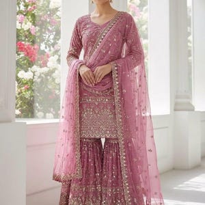 Op de afbeelding: Een mauvekleurig, geborduurd Sharara-pak met een bijpassende dupatta. De set heeft ingewikkelde gouden versieringen op het lijfje, de mouwen en de uitlopende broek. De dupatta is doorschijnend en ook versierd met gouden details. De outfit wordt gedragen met gouden oorbellen.