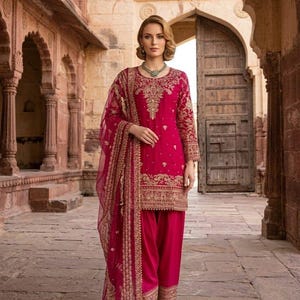 Peut inclure: Une femme porte un salwar kameez fuchsia, une tenue traditionnelle d'Asie du Sud. L'ensemble pr&eacute;sente des broderies dor&eacute;es complexes sur la tunique, le pantalon et le dupatta transparent. La tenue est compl&eacute;t&eacute;e par des chaussures assorties et un collier vert. En arri&egrave;re-plan, un b&acirc;timent en pierre.