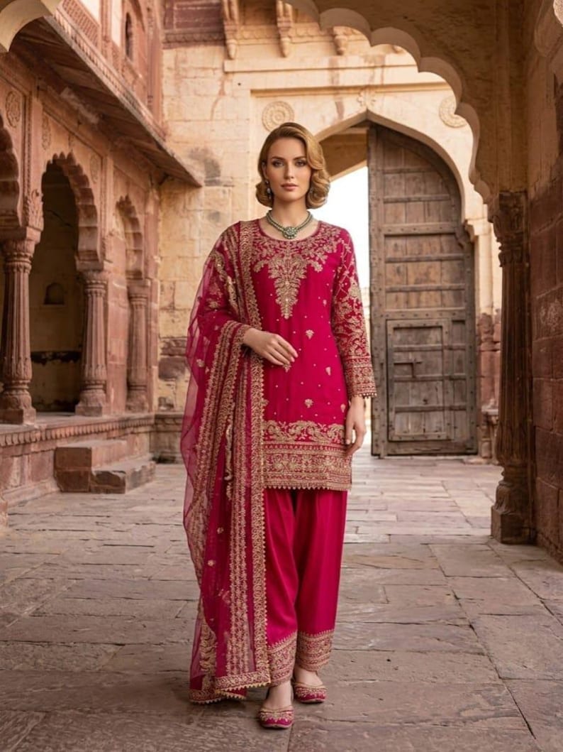 Peut inclure: Une femme porte un salwar kameez fuchsia, une tenue traditionnelle d'Asie du Sud. L'ensemble pr&eacute;sente des broderies dor&eacute;es complexes sur la tunique, le pantalon et le dupatta transparent. La tenue est compl&eacute;t&eacute;e par des chaussures assorties et un collier vert. En arri&egrave;re-plan, un b&acirc;timent en pierre.