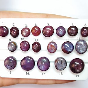 Puede incluir: Una colección de diecinueve piedras preciosas pulidas, cada una con forma de cabujón redondeado. Las piedras varían en color, desde rojos y púrpuras intensos hasta tonos más claros con patrones internos. Las piedras preciosas están dispuestas sobre una pantalla rectangular blanca.