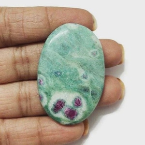 Puede incluir: Gema ovalada con una superficie verde moteada y pequeñas inclusiones rojas vibrantes. La piedra tiene una textura pulida y suave. La gema se sostiene en una mano, mostrando su belleza natural y variaciones de color únicas.