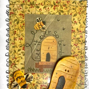 May include: A beige fabric with a floral pattern and the text "My Stitching Peace". The fabric is surrounded by a border of black and white buttons. There are two bee-shaped buttons and two beehive-shaped buttons on the fabric. The text "Bee ever so bumble" is written on the fabric.