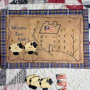 May include: A tan felt wall hanging with a blue and red plaid border. The center features an embroidered scene of a house with an American flag, two trees, and the text "Welcome two ewes". There are four white sheep buttons and three American flag buttons below the hanging.
