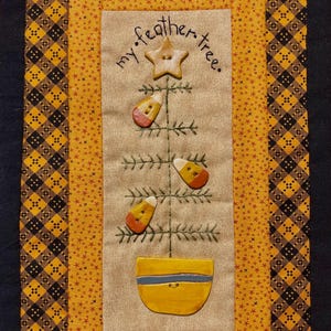 May include: A quilted wall hanging with a central panel featuring a stitched tree adorned with candy corn and a star. The text "my feather tree" is above the tree. The border has a yellow and black plaid pattern.