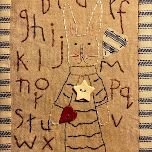 May include: A handmade fabric wall hanging featuring a stitched bunny wearing a striped shirt and holding a heart. The alphabet is embroidered around the bunny. The piece is framed with a blue and white striped fabric.