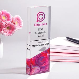 May include: A clear glass award with a pink and purple abstract design. The award is inscribed with "Channels ECO Leadership Award" and "Awarded to Madeline Porter".