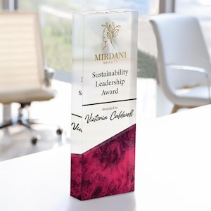 May include: A clear acrylic award with a white background and gold lettering. The award reads "Sustainability Leadership Award" and "Awarded to Victoria Caldwell". The award has a pink and white marbled design at the bottom.