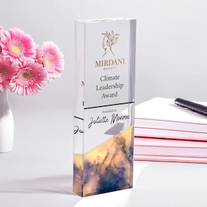 May include: A clear acrylic award with a gold and white logo that says "Mirdani Beauty". The award is for "Climate Leadership Award" and is awarded to "Juliette Monroe". The award has a watercolor design in shades of blue, yellow, and orange.