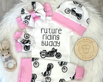 May include: A white baby outfit with a pink bow, hat, and leggings. The outfit features a black motorcycle design and the text "future riding buddy".