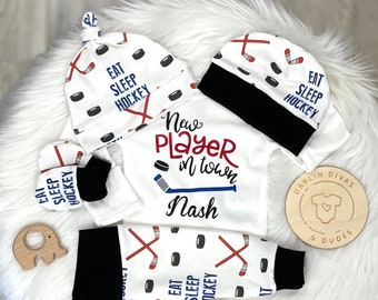 Pode incluir: Um conjunto de bebé branco com tema de hóquei. O conjunto inclui um chapéu, um macacão e leggings. O macacão tem o texto "Novo jogador na cidade" e o nome "Nash". O chapéu, o macacão e as leggings têm todos um design de taco de hóquei e disco com o texto "Comer Dormir Hóquei".