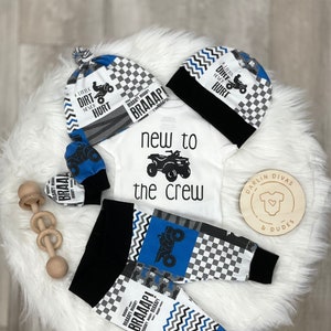 May include: A baby outfit set with a white onesie that says "new to the crew" with a blue ATV graphic, black and white checkered pants with blue and white zig-zag patterns, a blue and white hat with the text "A little dirt never hurt" and a blue and white mitten with the text "BRAAAP!"