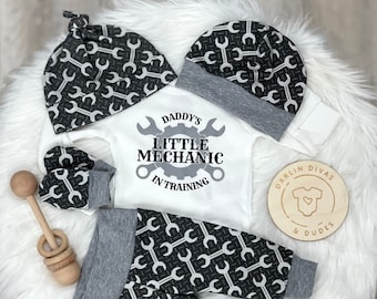 May include: A baby outfit set with a white onesie that says "Daddy's Little Mechanic in Training" with a wrench design, a gray and black beanie with a wrench pattern, gray and black pants with a wrench pattern, and a pair of gray and black mittens with a wrench pattern.