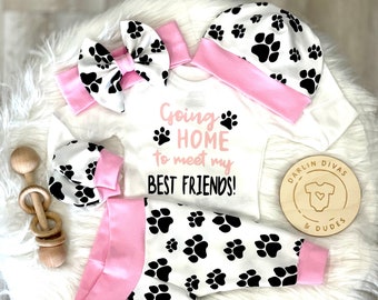 May include: A baby outfit set with a white onesie, pink and white hat, pink and white headband, white and black paw print leggings, and a wooden sign that says "Darlin' Divas & Dudes". The onesie has the text "Going Home to meet my Best Friends!" with paw prints.