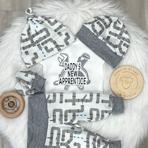 May include: A baby outfit set with a white bodysuit, gray pants, and a gray and white hat. The bodysuit has a graphic of a hard hat and tools with the text "Daddy's New Apprentice". The pants and hat have a repeating pattern of gray pipes on a white background.