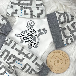 May include: A white baby onesie with the text "Daddy's New Apprentice" and a wrench and pipe design. The onesie is paired with a gray and white hat and pants with a gray and white pipe design. The hat and pants have a gray trim. A wooden circle with the text "Darlin Divas & Dudes" is in the lower right corner.