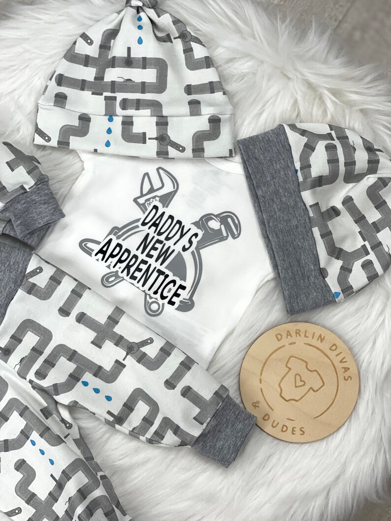 May include: A white baby onesie with the text "Daddy's New Apprentice" and a wrench and pipe design. The onesie is paired with a gray and white hat and pants with a gray and white pipe design. The hat and pants have a gray trim. A wooden circle with the text "Darlin Divas & Dudes" is in the lower right corner.