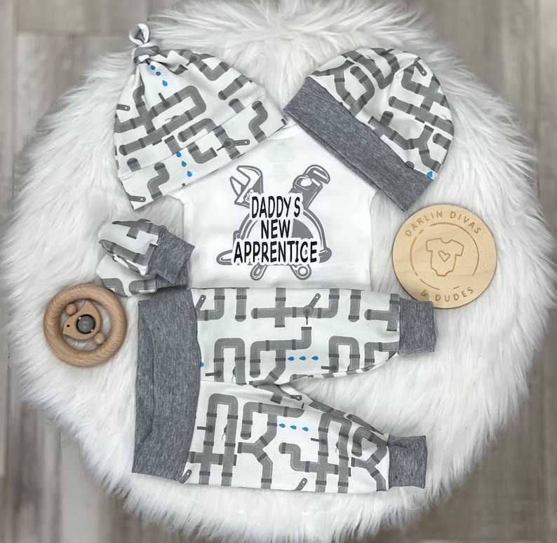 May include: A baby outfit set with a white bodysuit, gray pants, and a gray and white hat. The bodysuit has a graphic of a hard hat and tools with the text "Daddy's New Apprentice". The pants and hat have a repeating pattern of gray pipes on a white background.