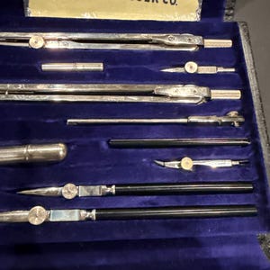 May include: A vintage drafting instrument set in a blue velvet-lined case. The set includes silver-toned metal compasses, dividers, and ruling pens with black handles. A label at the top reads YOUR INSTRUMENTS KEUFFEL & ESSER CO.
