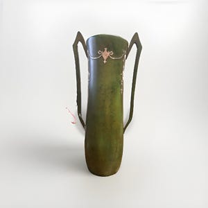 May include: A tall, cylindrical green vase with two elongated, angular handles extending from the sides. The surface features a mottled, weathered finish with a delicate, light-coloured decorative garland pattern encircling the upper section.