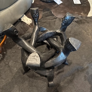 Puede incluir: Una escultura circular de madera en gris oscuro que presenta cuatro figuras estilizadas unidas en un anillo. Cada figura tiene una cabeza y un torso tallados y lisos, con los brazos extendidos para conectar con la siguiente, formando una estructura circular continua e interconectada.