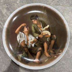 Può includere: Un piatto decorativo in ceramica con un dipinto classico di due giovani che condividono il cibo. L'opera raffigura un ragazzo che mangia uva e un altro che tiene in mano del pane, su uno sfondo marrone con un bordo bianco.