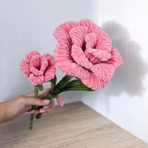Puede incluir: Dos flores rosas de pana con tallos verdes se sostienen sobre un fondo blanco. La flor más grande está en plena floración, mientras que la más pequeña está parcialmente abierta. Las flores están hechas de un material suave y texturizado.