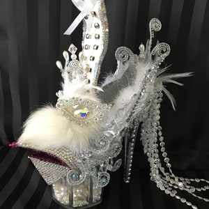 May include: A clear high-heeled shoe with a white fur pom-pom, a crystal crown, and a crystal beaded fringe. The shoe is decorated with rhinestones and pearls.