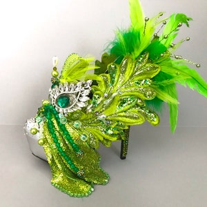 May include: A green and silver high heel shoe with a large green jewel and feathers. The shoe is decorated with sequins and beads.