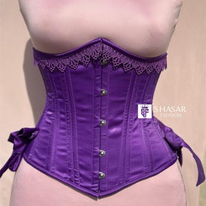 May include: A vibrant purple corset with a satin finish. The corset features a delicate lace trim along the top edge and is adorned with silver-toned clasps down the front. Bows are tied on each side. The corset is displayed on a mannequin.