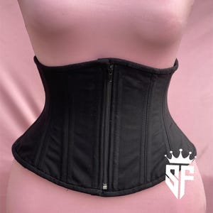 May include: Black corset with a front zipper closure. The corset has vertical seams and a curved shape, designed to cinch the waist. The garment is displayed on a mannequin against a pink background.