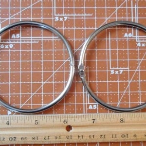 May include: Two silver-colored metal binder rings are displayed on a brown cutting mat with grid lines and a wooden ruler. The rings are circular with a hinged opening. The ruler shows inch measurements.