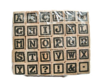 May include: A set of wooden alphabet blocks, each with a black letter or symbol printed on a square face. The blocks are packaged in clear plastic, and the letters are in a bold, sans-serif font. The set includes letters A through Z, plus punctuation.