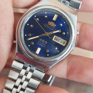 Op de afbeelding: Een zilverkleurig Orient horloge met een donkerblauwe wijzerplaat. Het horloge heeft goudkleurige uurmarkeringen en wijzers, een dag-datumweergave en een roestvrijstalen band. De wijzerplaat leest "Orient Crystal 21 Jewels".