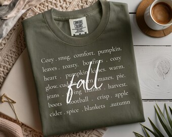 May include: Olive green Comfort Colors t-shirt with the word "fall" in white script. The shirt features a list of fall-themed words like "pumpkin," "cozy," and "apple." A cup of coffee sits on a wooden coaster in the background.