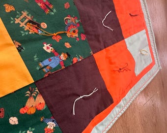 May include: A patchwork quilt with a green, orange, brown, and yellow color scheme. The quilt is made of different fabrics with various patterns, including a pattern of scarecrows and pumpkins. The quilt has a light blue border with a decorative stitch.