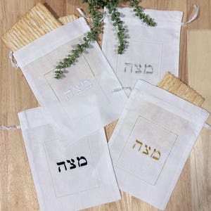May include: Four white linen drawstring bags with the Hebrew word "Matzah" embroidered in different colors: white, silver, black, and gold. Each bag has a rectangular embroidered border. Matzah crackers are visible in the background.
